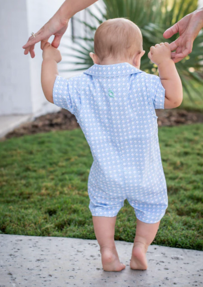 A baby in a blue Quail Blue Cane Lattice Guayabera Romper walks on grass with an adult’s support, set against lush greenery and a white wall in the background.
