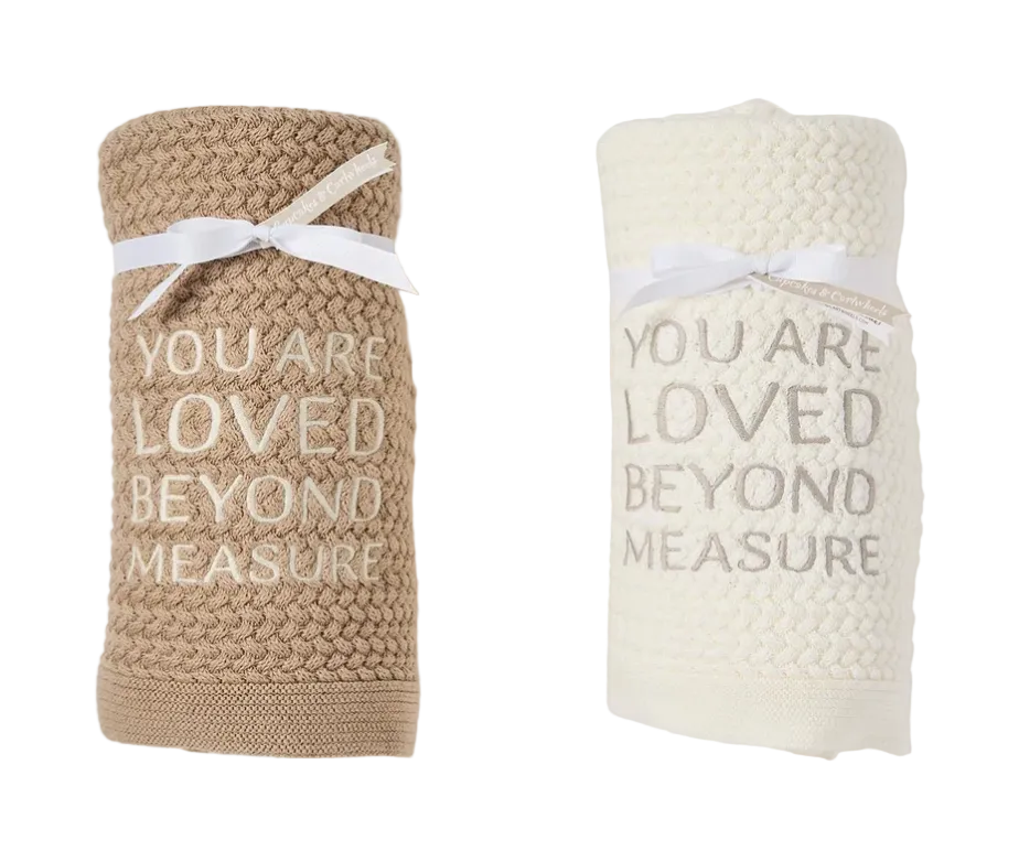 Two rolled knit baby blankets on a white background, one in warm beige with white text reading 'YOU ARE LOVED BEYOND MEASURE' tied with a white ribbon, and one in off-white with taupe text reading 'YOU ARE LOVED BEYOND MEASURE' tied with a white ribbon.
