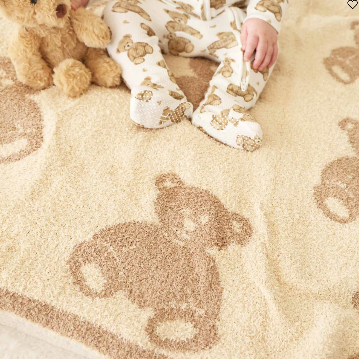 A close-up of a plush cream-colored blanket with a brown teddy bear pattern, with a baby's legs and a stuffed teddy bear visible at the top edge.