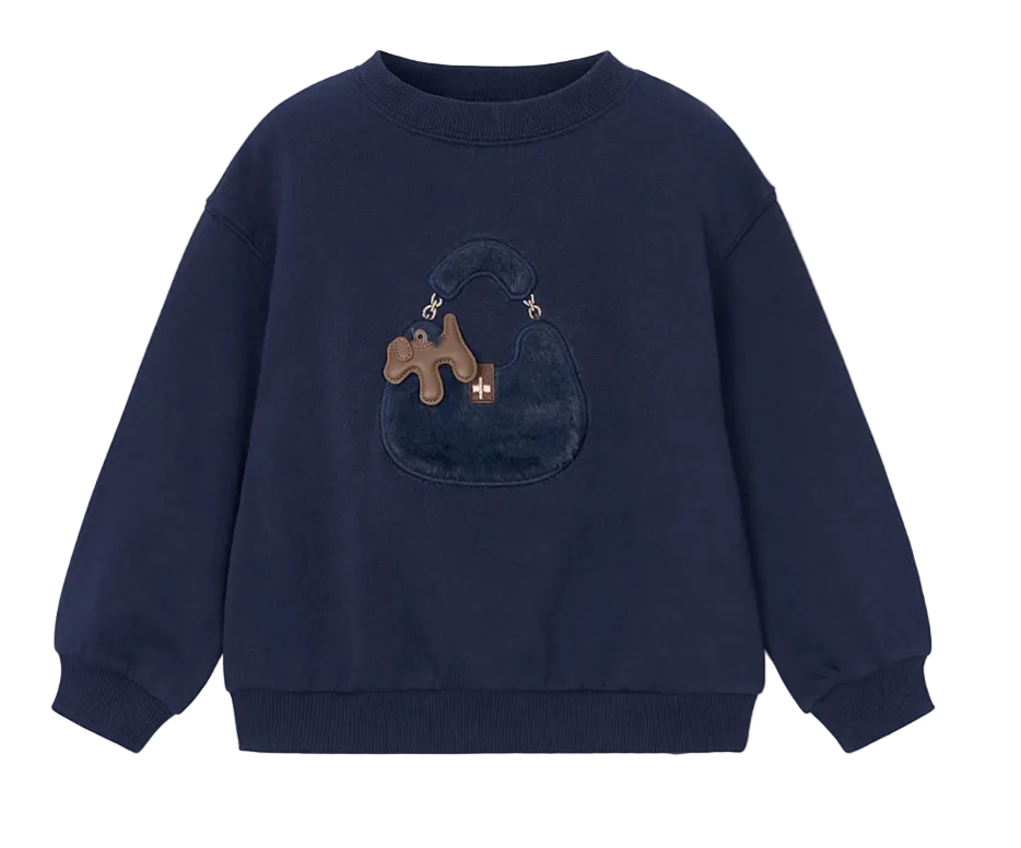 A navy blue crewneck sweatshirt featuring a plush, textured applique of a dark blue handbag with a brown puzzle-shaped charm on the front, displayed against a light gray background.