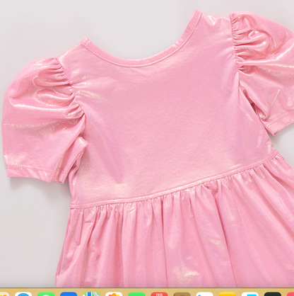 The Light Pink Laurie Dress by Pink Chicken features shiny lamé fabric, puffed short sleeves, and a gathered waist, displayed flat on a light surface.
