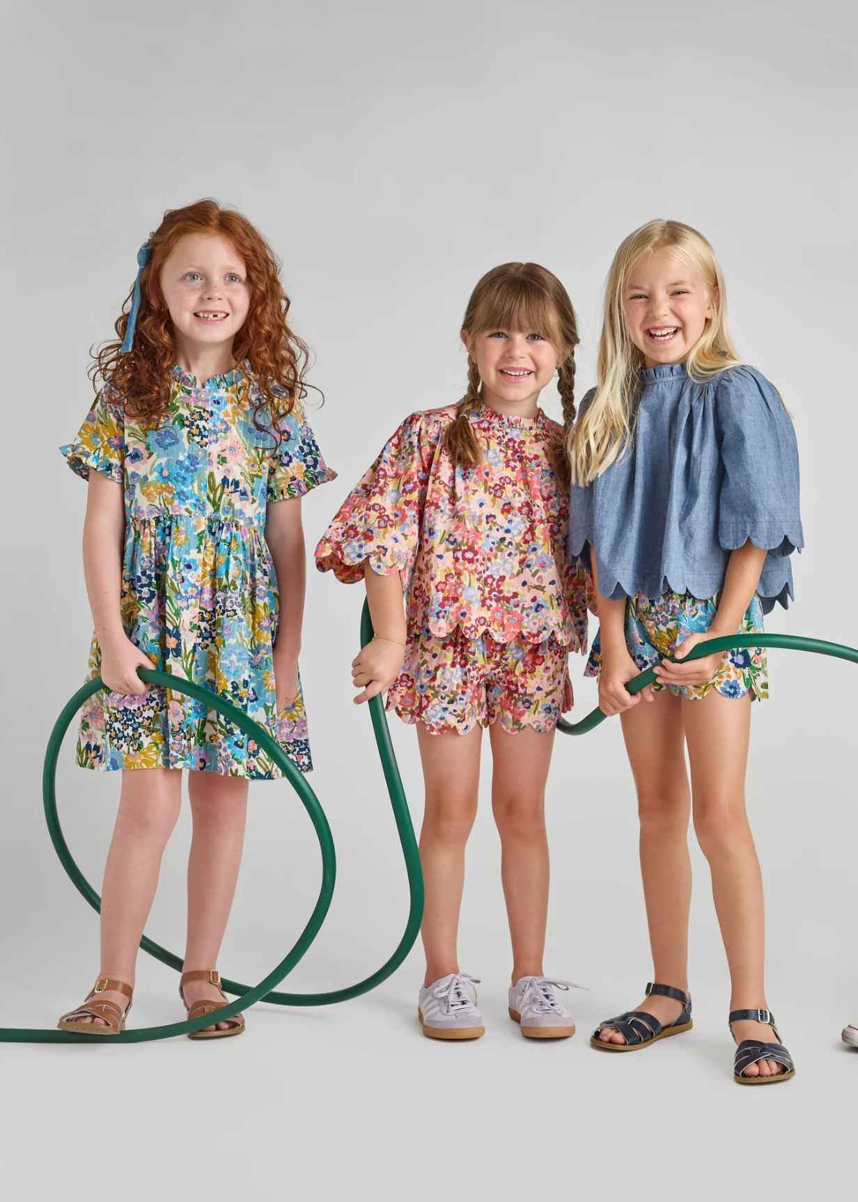 Three young girls smile with garden hoses against a plain background. One wears the Pink Chicken Sketch Floral Autumn Dress, another a floral top and shorts, and the third a blue ruffled top with printed shorts.