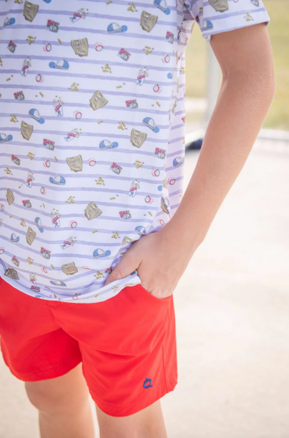 A person, outdoors on a sunny day, wears the blue Quail 7th Inning Polo—white with light blue stripes and playful colorful patterns—paired with bright red shorts, standing with one hand in their pocket.