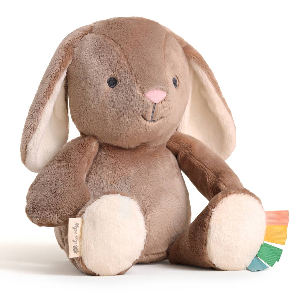 The Itzy Ritzy Sweetie Snuggles™ Plush is a soft brown bunny with cream paws and ears, a pink nose, stitched smile, and colorful ribbon tags on one paw for sensory play. This cuddly plush sits upright on a white background.
