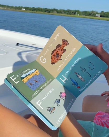 Someone sits on a boat holding ABC's of Fishing by Wild Adventure Books, open to pages E-H. Water, greenery, and a clear blue sky make it an ideal fishing gift scene for kids.